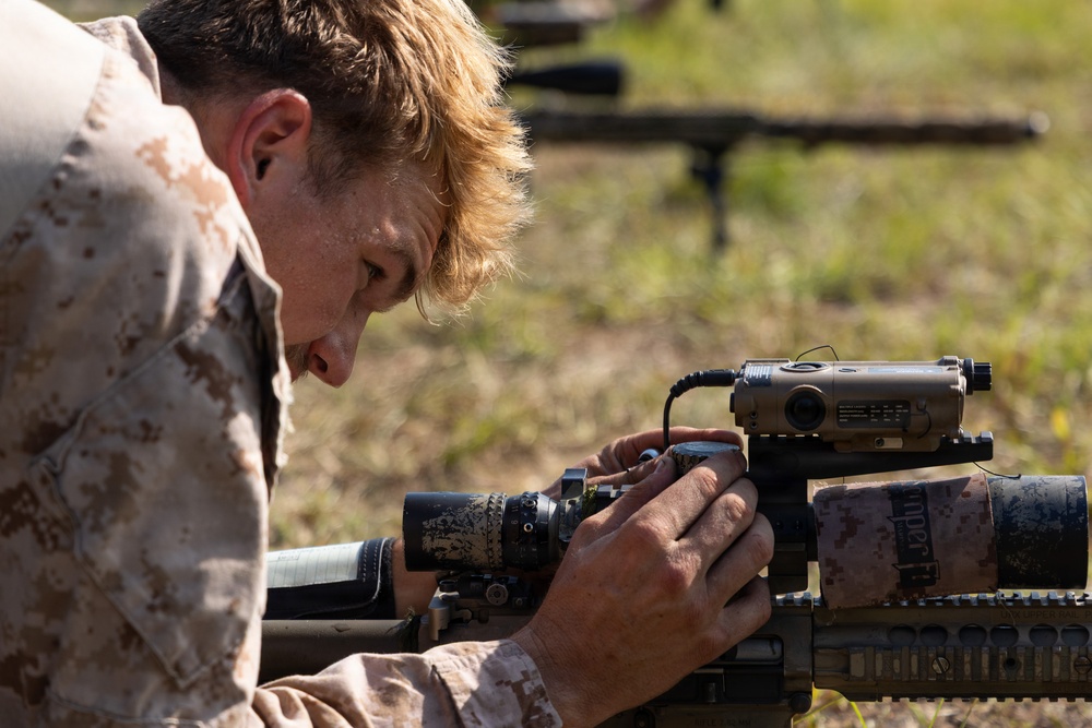 A military personnel wearing desert MARPAT camouflage uniform is closely inspecting or adjusting optical/electronic equipment mounted on a precision rifle. The individual appears to be on a shooting range, prone or leaning over the weapon system. Additional rifle systems on bipods are visible in the blurred background.