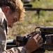 A military personnel wearing desert MARPAT camouflage uniform is closely inspecting or adjusting optical/electronic equipment mounted on a precision rifle. The individual appears to be on a shooting range, prone or leaning over the weapon system. Additional rifle systems on bipods are visible in the blurred background.