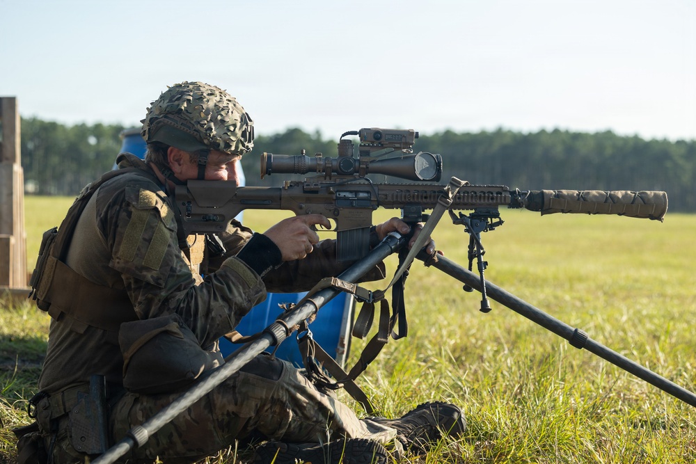 A military or law enforcement operator is positioned in a prone/seated firing position on a grass field, aiming a large precision rifle mounted on a bipod. The shooter is equipped with full tactical gear including a camouflage-covered helmet, body armor, and a plate carrier with pouches. The setting appears to be an outdoor shooting range or training exercise.