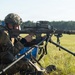 A military or law enforcement operator is positioned in a prone/seated firing position on a grass field, aiming a large precision rifle mounted on a bipod. The shooter is equipped with full tactical gear including a camouflage-covered helmet, body armor, and a plate carrier with pouches. The setting appears to be an outdoor shooting range or training exercise.