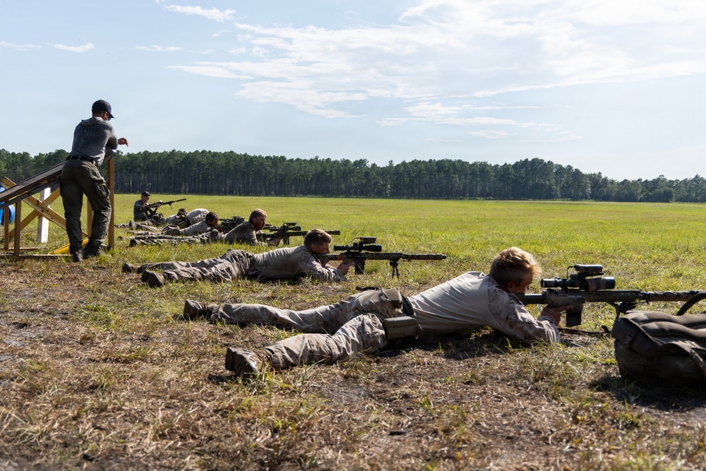 A group of shooters are in prone positions on a grass firing line, aiming long-range precision rifles downrange across an open field. An instructor or range officer stands on an elevated platform to the left, observing the shooters. The setting appears to be an outdoor shooting range or military/law enforcement training exercise.