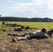 A group of shooters are in prone positions on a grass firing line, aiming long-range precision rifles downrange across an open field. An instructor or range officer stands on an elevated platform to the left, observing the shooters. The setting appears to be an outdoor shooting range or military/law enforcement training exercise.