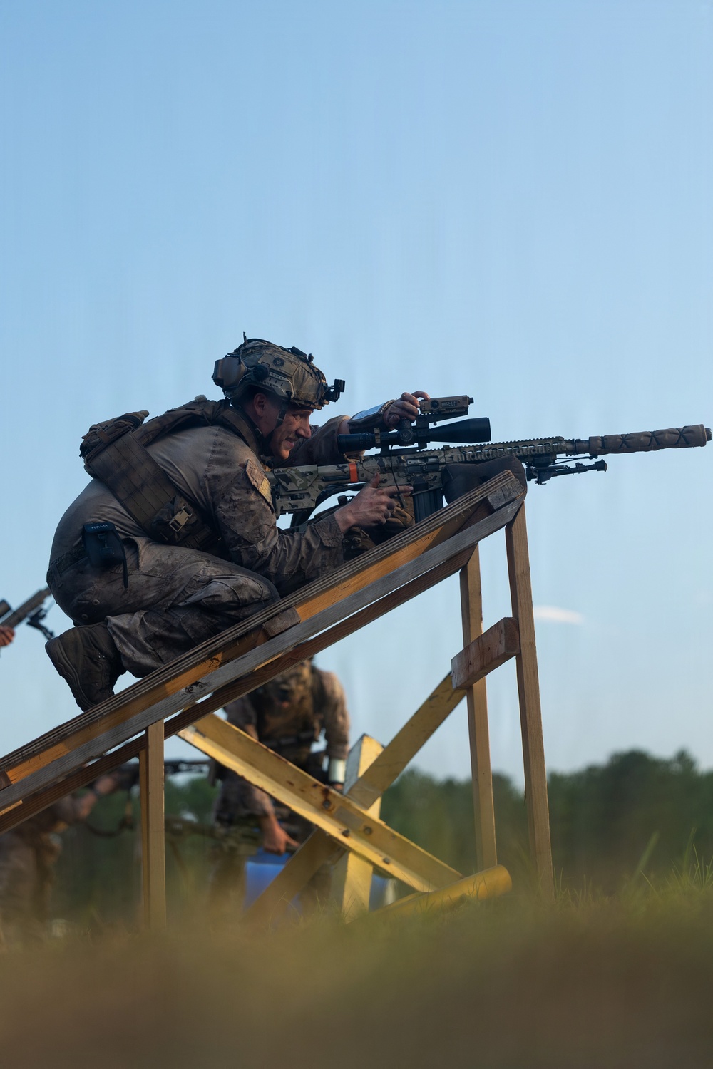 A heavily equipped soldier in multicam/OCP camouflage uniform is positioned on a wooden elevated platform, aiming a scoped rifle with a suppressor attached. The soldier is wearing a tactical helmet with night-vision device mount, hearing protection, and a full plate carrier with accessories. A second soldier in similar gear is visible in the background at a lower position.