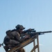 A heavily equipped soldier in multicam/OCP camouflage uniform is positioned on a wooden elevated platform, aiming a scoped rifle with a suppressor attached. The soldier is wearing a tactical helmet with night-vision device mount, hearing protection, and a full plate carrier with accessories. A second soldier in similar gear is visible in the background at a lower position.