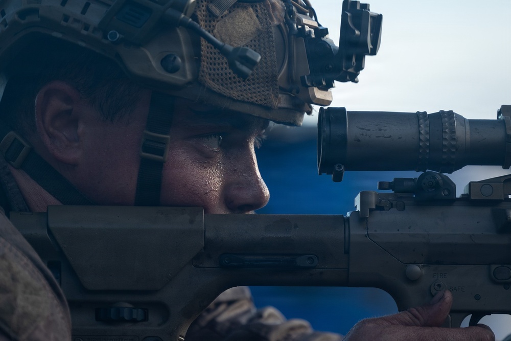 A military service member is shown in close-up profile view, aiming down the scope of a precision rifle. The individual is equipped with tactical gear including a combat helmet with night vision device mount and communications equipment. The image captures a training or operational shooting posture with the weapon's fire selector switch visible.