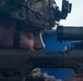 A military service member is shown in close-up profile view, aiming down the scope of a precision rifle. The individual is equipped with tactical gear including a combat helmet with night vision device mount and communications equipment. The image captures a training or operational shooting posture with the weapon's fire selector switch visible.