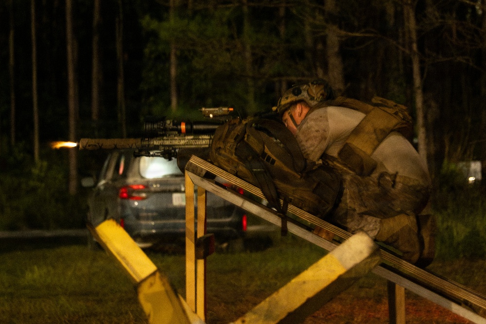 A heavily equipped tactical operator is photographed at night firing a rifle from behind a wooden barricade structure. A muzzle flash is visible at the end of the weapon's barrel, indicating the weapon is being discharged. The setting appears to be a training range or exercise area, with a parked vehicle and tree line visible in the background.