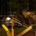 A heavily equipped tactical operator is photographed at night firing a rifle from behind a wooden barricade structure. A muzzle flash is visible at the end of the weapon's barrel, indicating the weapon is being discharged. The setting appears to be a training range or exercise area, with a parked vehicle and tree line visible in the background.