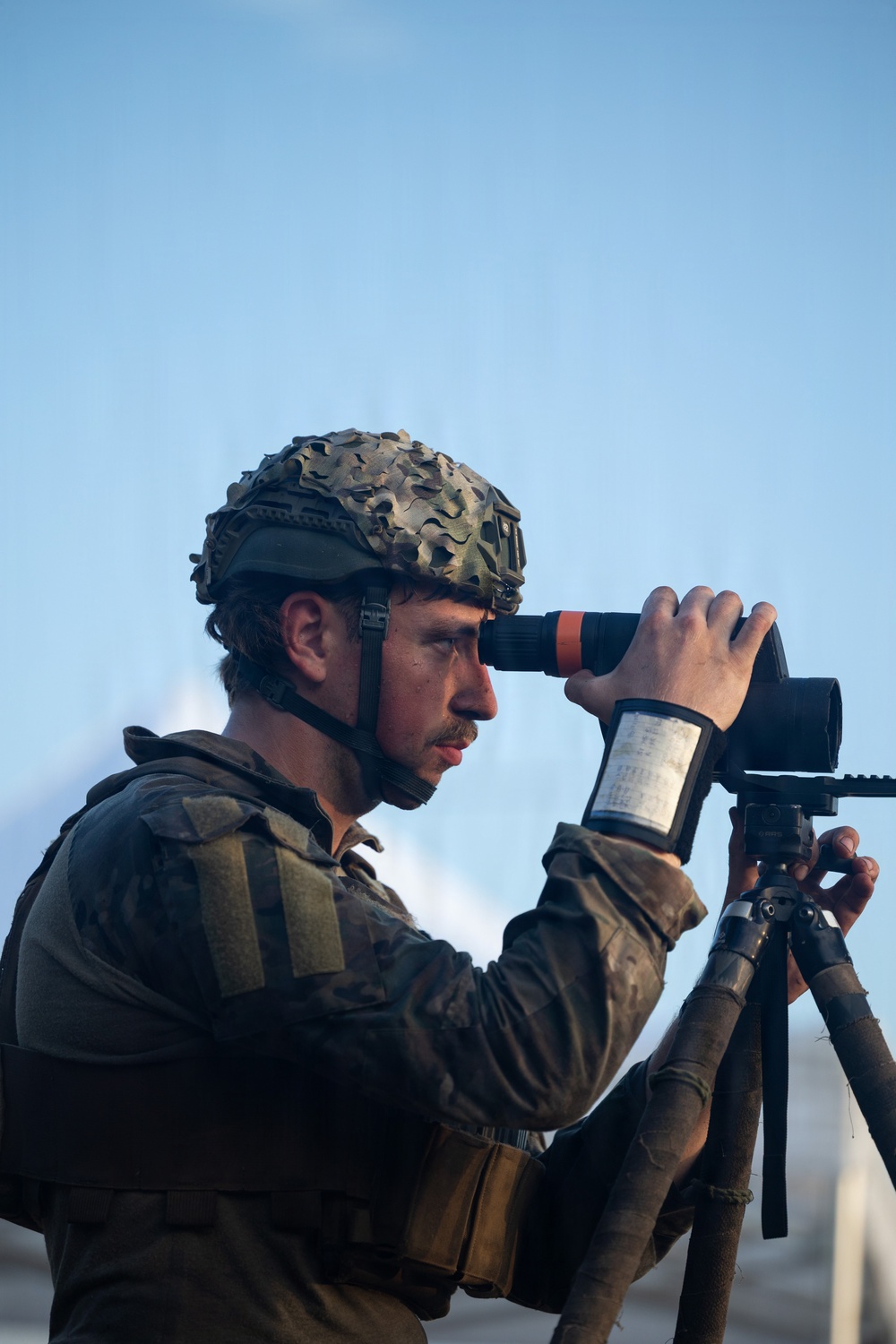 A soldier in military camouflage uniform and helmet with camouflage netting is using a tripod-mounted optical observation device, likely a thermal or digital spotting scope. The individual appears to be conducting surveillance or reconnaissance, looking through the eyepiece of the device. The image is taken against a clear blue sky background.