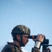 A soldier in military camouflage uniform and helmet with camouflage netting is using a tripod-mounted optical observation device, likely a thermal or digital spotting scope. The individual appears to be conducting surveillance or reconnaissance, looking through the eyepiece of the device. The image is taken against a clear blue sky background.