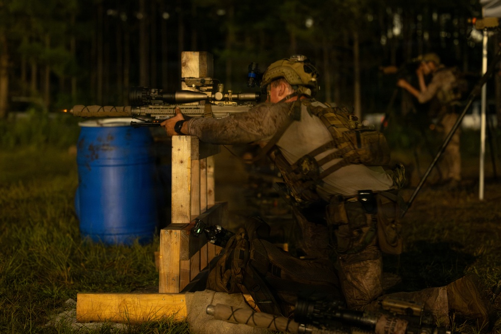 A military personnel in full combat gear is positioned behind a wooden barricade, aiming a scoped rifle in a low-light training environment. The setting appears to be an outdoor range or tactical training exercise, with a blue barrel and woodland backdrop visible. A second individual in the background appears to be observing or documenting the exercise.