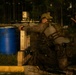 A military personnel in full combat gear is positioned behind a wooden barricade, aiming a scoped rifle in a low-light training environment. The setting appears to be an outdoor range or tactical training exercise, with a blue barrel and woodland backdrop visible. A second individual in the background appears to be observing or documenting the exercise.