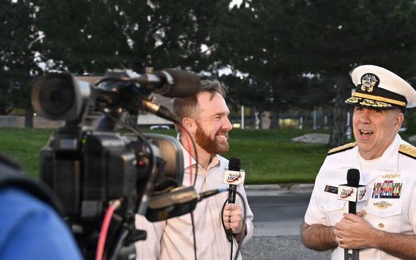 A U.S. Navy flag officer in white dress uniform is being interviewed by a television reporter holding a branded microphone, while a camera operator records the exchange. The setting appears to be an outdoor location, likely a military installation or public venue. Both the officer and reporter appear engaged and smiling during the interview.