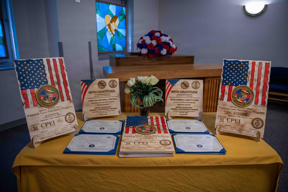 A ceremonial display table covered with a gold/yellow tablecloth showcasing awards and certificates for the United States Navy Pastoral Care Residency Program. The table features wooden plaques with American flag designs, 'With Gratitude' recognition plaques, CPEI certificates, and a central floral arrangement. The setting appears to be a chapel or religious space, indicated by the stained glass window and altar visible in the background.