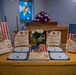 A ceremonial display table covered with a gold/yellow tablecloth showcasing awards and certificates for the United States Navy Pastoral Care Residency Program. The table features wooden plaques with American flag designs, 'With Gratitude' recognition plaques, CPEI certificates, and a central floral arrangement. The setting appears to be a chapel or religious space, indicated by the stained glass window and altar visible in the background.