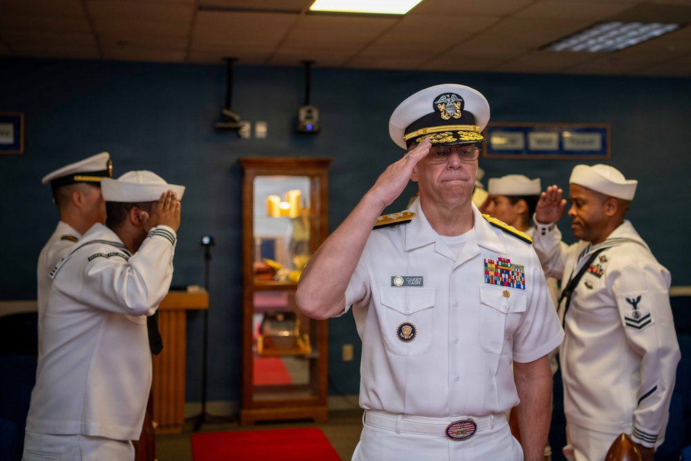 A U.S. Navy officer of senior rank, wearing a white service dress uniform with multiple ribbons and gold shoulder boards, renders a salute during what appears to be a formal military ceremony. Several enlisted sailors in white uniforms are also saluting in the background. The setting appears to be an indoor military facility or command room.