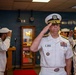 A U.S. Navy officer of senior rank, wearing a white service dress uniform with multiple ribbons and gold shoulder boards, renders a salute during what appears to be a formal military ceremony. Several enlisted sailors in white uniforms are also saluting in the background. The setting appears to be an indoor military facility or command room.