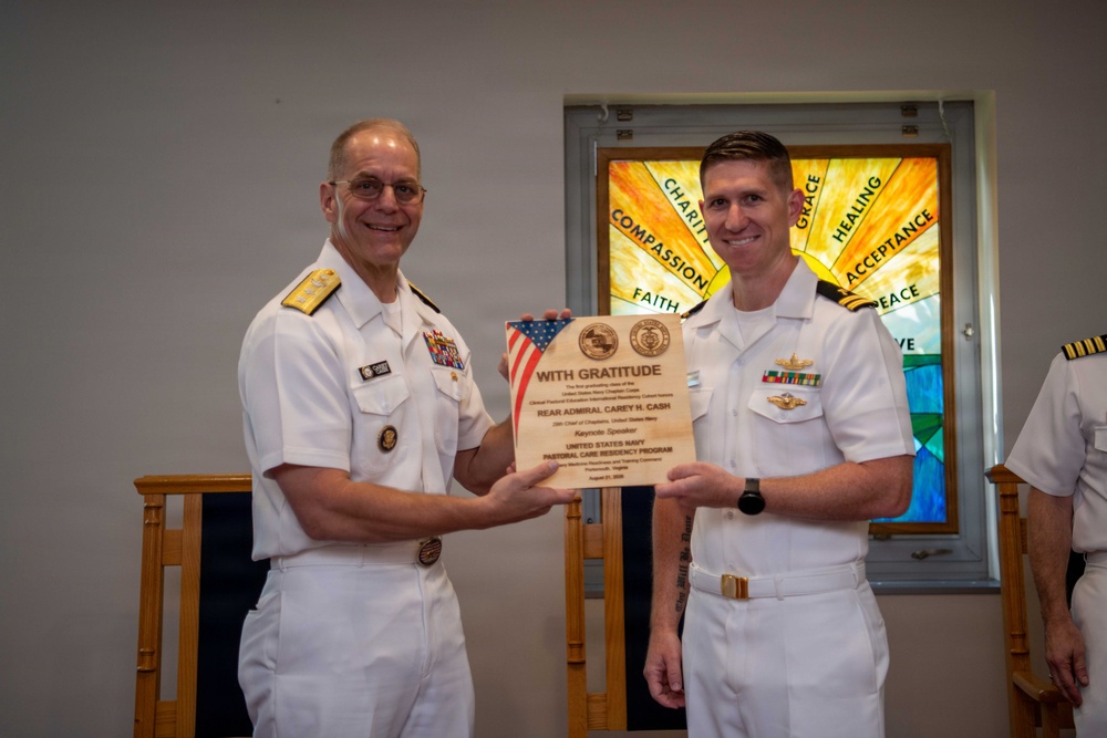 Two U.S. Navy officers in white dress uniforms are posing together while holding a wooden plaque inscribed 'With Gratitude' recognizing Rear Admiral Carey H. Cash as a keynote speaker for the United States Navy Pastoral Care Residency Program. The ceremony appears to take place in a chapel setting, evidenced by the stained glass window visible in the background displaying words such as 'Charity,' 'Compassion,' 'Faith,' 'Grace,' 'Healing,' 'Acceptance,' and 'Peace.' A third uniformed officer is partially visible on the right edge of the frame.