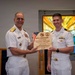 Two U.S. Navy officers in white dress uniforms are posing together while holding a wooden plaque inscribed 'With Gratitude' recognizing Rear Admiral Carey H. Cash as a keynote speaker for the United States Navy Pastoral Care Residency Program. The ceremony appears to take place in a chapel setting, evidenced by the stained glass window visible in the background displaying words such as 'Charity,' 'Compassion,' 'Faith,' 'Grace,' 'Healing,' 'Acceptance,' and 'Peace.' A third uniformed officer is partially visible on the right edge of the frame.
