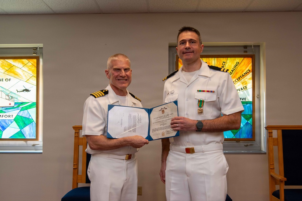 Two U.S. Navy officers in white dress uniforms are posing together during a formal award ceremony. The officer on the right is wearing a newly pinned medal on his chest, while both hold open a Department of the Navy certificate. The setting appears to be a chapel or religious facility, evidenced by stained glass windows visible in the background.