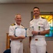 Two U.S. Navy officers in white dress uniforms are posing together during a formal award ceremony. The officer on the right is wearing a newly pinned medal on his chest, while both hold open a Department of the Navy certificate. The setting appears to be a chapel or religious facility, evidenced by stained glass windows visible in the background.