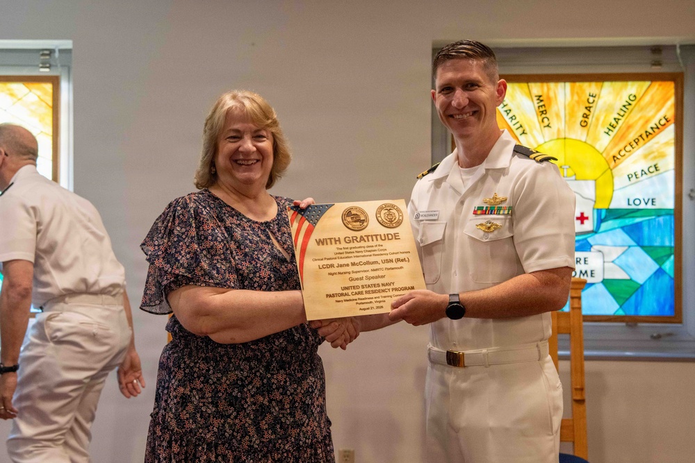 A woman and a U.S. Navy officer in dress whites are jointly holding a wooden plaque that reads 'With Gratitude' from the United States Navy Pastoral Care Residency Program. The plaque is being presented to LCDR Jane McCollum, USN (Ret.), recognized as a Guest Speaker. The ceremony appears to be taking place in a chapel or religious setting, evidenced by stained glass windows in the background.