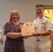 A woman and a U.S. Navy officer in dress whites are jointly holding a wooden plaque that reads 'With Gratitude' from the United States Navy Pastoral Care Residency Program. The plaque is being presented to LCDR Jane McCollum, USN (Ret.), recognized as a Guest Speaker. The ceremony appears to be taking place in a chapel or religious setting, evidenced by stained glass windows in the background.
