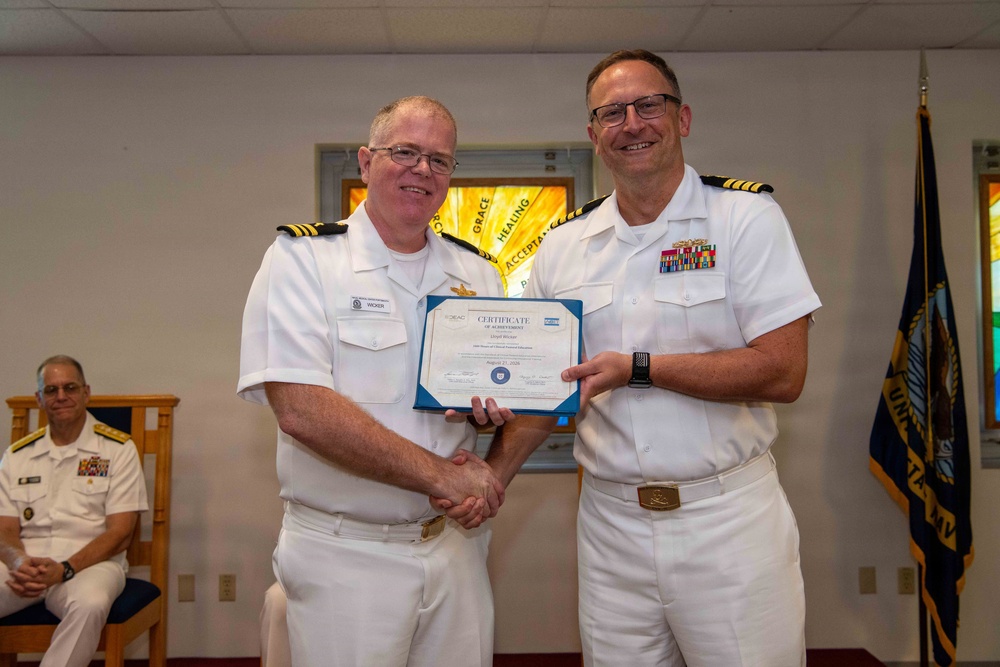 Two naval officers in white dress uniforms are shaking hands while jointly holding a Certificate of Achievement during a formal military ceremony. A third officer is seated in the background observing the presentation. The setting appears to be a chapel or meeting room with stained glass visible behind them.