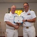 Two naval officers in white dress uniforms are shaking hands while jointly holding a Certificate of Achievement during a formal military ceremony. A third officer is seated in the background observing the presentation. The setting appears to be a chapel or meeting room with stained glass visible behind them.