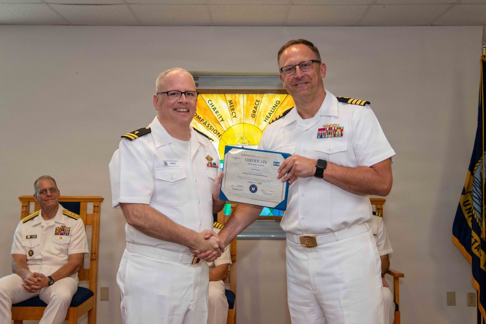 Two U.S. Navy officers in white dress uniforms are shaking hands while one presents a Certificate of Achievement to the other. The ceremony appears to take place in a chapel setting, evidenced by a stained glass window in the background displaying words such as 'Charity,' 'Mercy,' 'Grace,' and 'Healing.' A third officer is seated in the background observing the presentation.