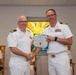 Two U.S. Navy officers in white dress uniforms are shaking hands while one presents a Certificate of Achievement to the other. The ceremony appears to take place in a chapel setting, evidenced by a stained glass window in the background displaying words such as 'Charity,' 'Mercy,' 'Grace,' and 'Healing.' A third officer is seated in the background observing the presentation.