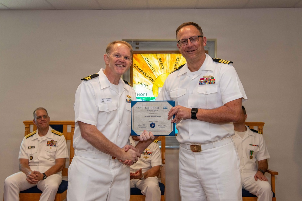 Two U.S. Navy officers in white dress uniforms are shaking hands while jointly holding a Certificate of Achievement during a formal military ceremony. The officer on the right is presenting the certificate to the officer on the left, both smiling for the photo. Several other uniformed military personnel are seated in the background observing the ceremony.