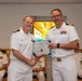 Two U.S. Navy officers in white dress uniforms are shaking hands while jointly holding a Certificate of Achievement during a formal military ceremony. The officer on the right is presenting the certificate to the officer on the left, both smiling for the photo. Several other uniformed military personnel are seated in the background observing the ceremony.