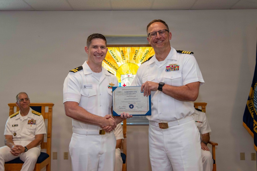 Two U.S. Navy officers in white dress uniforms are shaking hands while jointly holding a Certificate of Achievement during a formal military ceremony. The certificate appears to be from BUMED (Bureau of Medicine and Surgery) and is dated August 21, 2024. A third officer is seated in the background observing the ceremony, which takes place in what appears to be a chapel or ceremonial room.