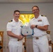 Two U.S. Navy officers in white dress uniforms are shaking hands while jointly holding a Certificate of Achievement during a formal military ceremony. The certificate appears to be from BUMED (Bureau of Medicine and Surgery) and is dated August 21, 2024. A third officer is seated in the background observing the ceremony, which takes place in what appears to be a chapel or ceremonial room.