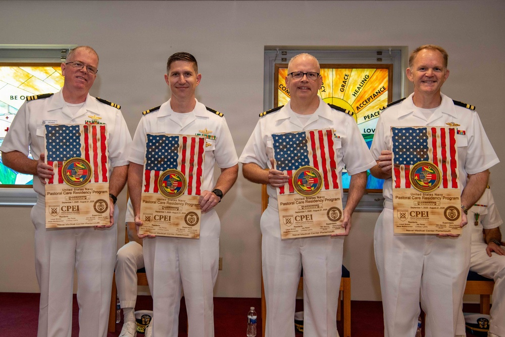 Four U.S. Navy officers in white dress uniforms are posing together while holding wooden plaques featuring the American flag and an official seal. The plaques appear to be certificates from the United States Navy Pastoral Care Residency Program, issued by Naval Medical Center Portsmouth. The setting appears to be a chapel or religious space, indicated by stained glass windows visible in the background.