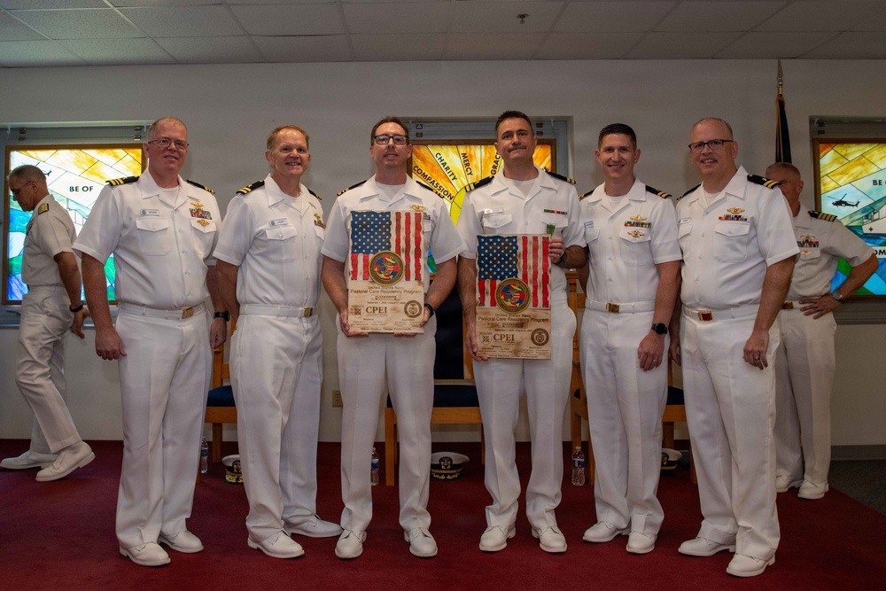 Six U.S. Navy personnel in white dress uniforms pose for a group photo in what appears to be a chapel or religious facility. Two of the individuals are holding wooden plaques featuring American flags and CPEI (Pastoral Care Resiliency Program) insignia. The setting suggests a formal military ceremony or award presentation.