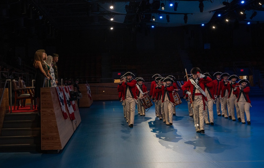 A ceremonial military fife and drum corps dressed in 18th-century Continental Army-style uniforms — red coats, white breeches, and tricorn hats — marches across a blue-lit indoor performance floor. The performers pass a raised reviewing stand where several civilians and at least one uniformed military officer are observing. The setting appears to be a formal military ceremony or tattoo event held in an indoor arena or auditorium.