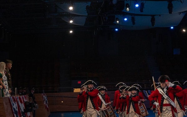 A ceremonial military fife and drum corps dressed in 18th-century Continental Army-style uniforms — red coats, white breeches, and tricorn hats — marches across a blue-lit indoor performance floor. The performers pass a raised reviewing stand where several civilians and at least one uniformed military officer are observing. The setting appears to be a formal military ceremony or tattoo event held in an indoor arena or auditorium.