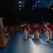 A ceremonial military fife and drum corps dressed in 18th-century Continental Army-style uniforms — red coats, white breeches, and tricorn hats — marches across a blue-lit indoor performance floor. The performers pass a raised reviewing stand where several civilians and at least one uniformed military officer are observing. The setting appears to be a formal military ceremony or tattoo event held in an indoor arena or auditorium.