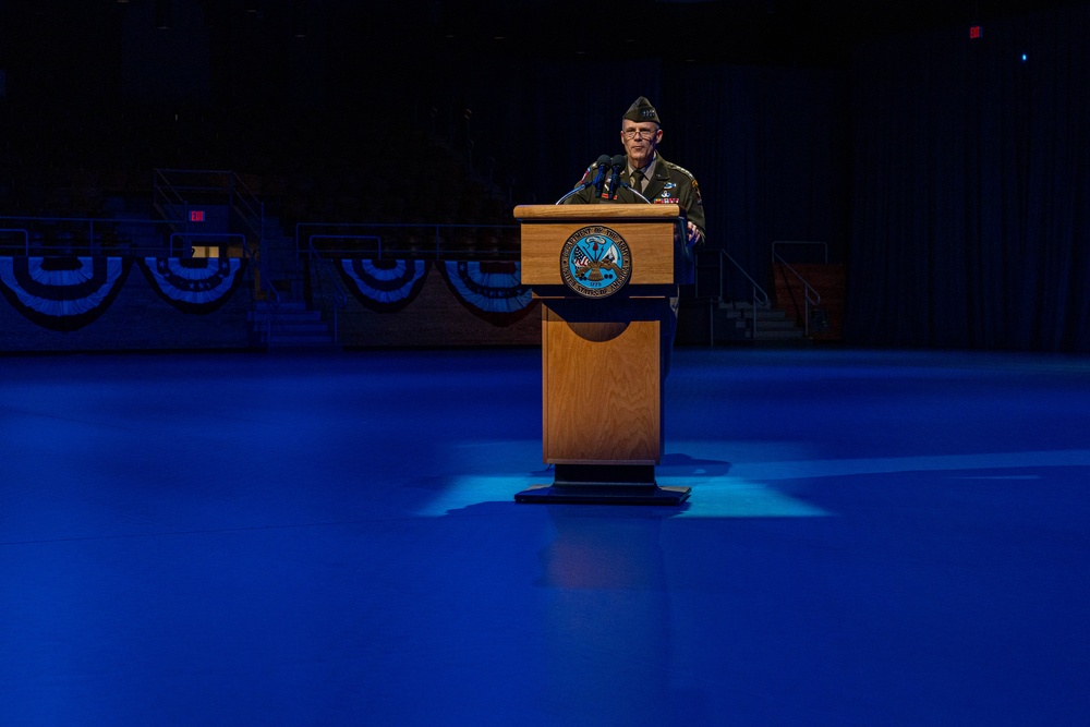 A military officer in dress uniform stands at a wooden podium on a stage illuminated by blue lighting. The setting appears to be a formal military ceremony or official event, with patriotic bunting decorations visible in the background. The podium bears what appears to be a Department of Defense or military branch seal.