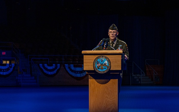 A military officer in dress uniform stands at a wooden podium on a stage illuminated by blue lighting. The setting appears to be a formal military ceremony or official event, with patriotic bunting decorations visible in the background. The podium bears what appears to be a Department of Defense or military branch seal.