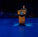 A military officer in dress uniform stands at a wooden podium on a stage illuminated by blue lighting. The setting appears to be a formal military ceremony or official event, with patriotic bunting decorations visible in the background. The podium bears what appears to be a Department of Defense or military branch seal.