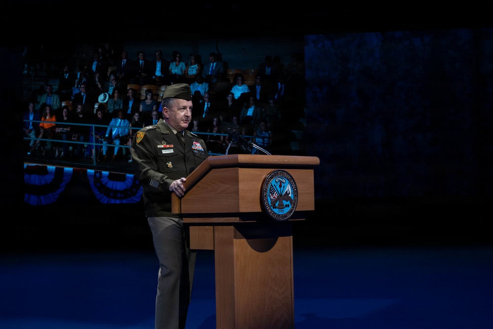 A U.S. Army officer in dress green uniform stands at a wooden podium delivering a speech at a formal military ceremony. The podium bears an official U.S. Army seal, and a large projection screen behind him displays an audience seated in tiered seating. The setting appears to be a large auditorium or arena with dramatic stage lighting.