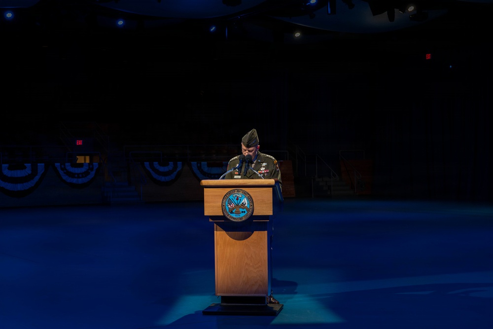 A military officer in dress uniform stands at a wooden podium on a darkened stage, appearing to deliver a speech or read remarks. The stage is decorated with blue and red bunting in the background, suggesting a formal military ceremony or official event. The scene is dramatically lit with blue stage lighting, isolating the speaker at the podium.