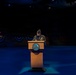 A military officer in dress uniform stands at a wooden podium on a darkened stage, appearing to deliver a speech or read remarks. The stage is decorated with blue and red bunting in the background, suggesting a formal military ceremony or official event. The scene is dramatically lit with blue stage lighting, isolating the speaker at the podium.