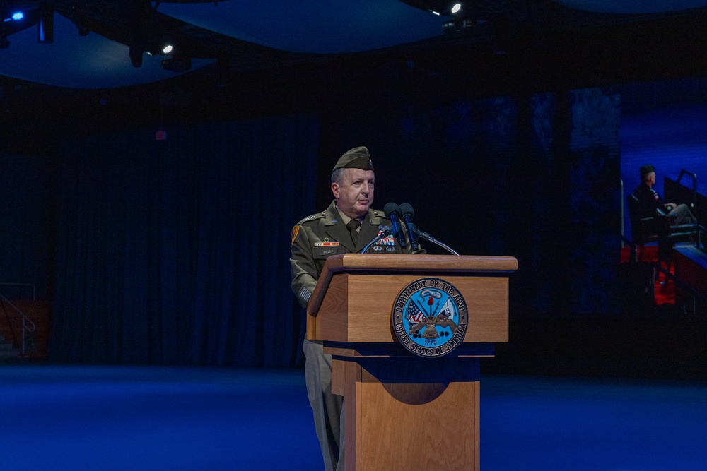 A U.S. Army soldier in dress uniform is speaking at a podium bearing the official seal of the Department of the Army. The event appears to be a formal military ceremony held in a large, darkened auditorium or theater with blue ambient lighting. The soldier is wearing a garrison cap and multiple medals/ribbons on their uniform.