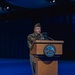 A U.S. Army soldier in dress uniform is speaking at a podium bearing the official seal of the Department of the Army. The event appears to be a formal military ceremony held in a large, darkened auditorium or theater with blue ambient lighting. The soldier is wearing a garrison cap and multiple medals/ribbons on their uniform.
