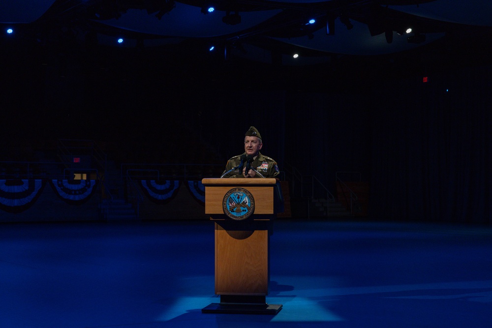 A military officer in camouflage uniform and garrison cap stands at a wooden podium bearing what appears to be a Department of Defense or U.S. Army seal. The setting is a darkened auditorium or theater stage illuminated by blue stage lighting, with patriotic bunting decorations visible in the background. The scene suggests a formal military ceremony or official address.