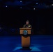 A military officer in camouflage uniform and garrison cap stands at a wooden podium bearing what appears to be a Department of Defense or U.S. Army seal. The setting is a darkened auditorium or theater stage illuminated by blue stage lighting, with patriotic bunting decorations visible in the background. The scene suggests a formal military ceremony or official address.